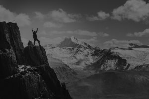 Man on top of a mountain with hands held high towards big puffy clouds and a wonderful view and blue skies.